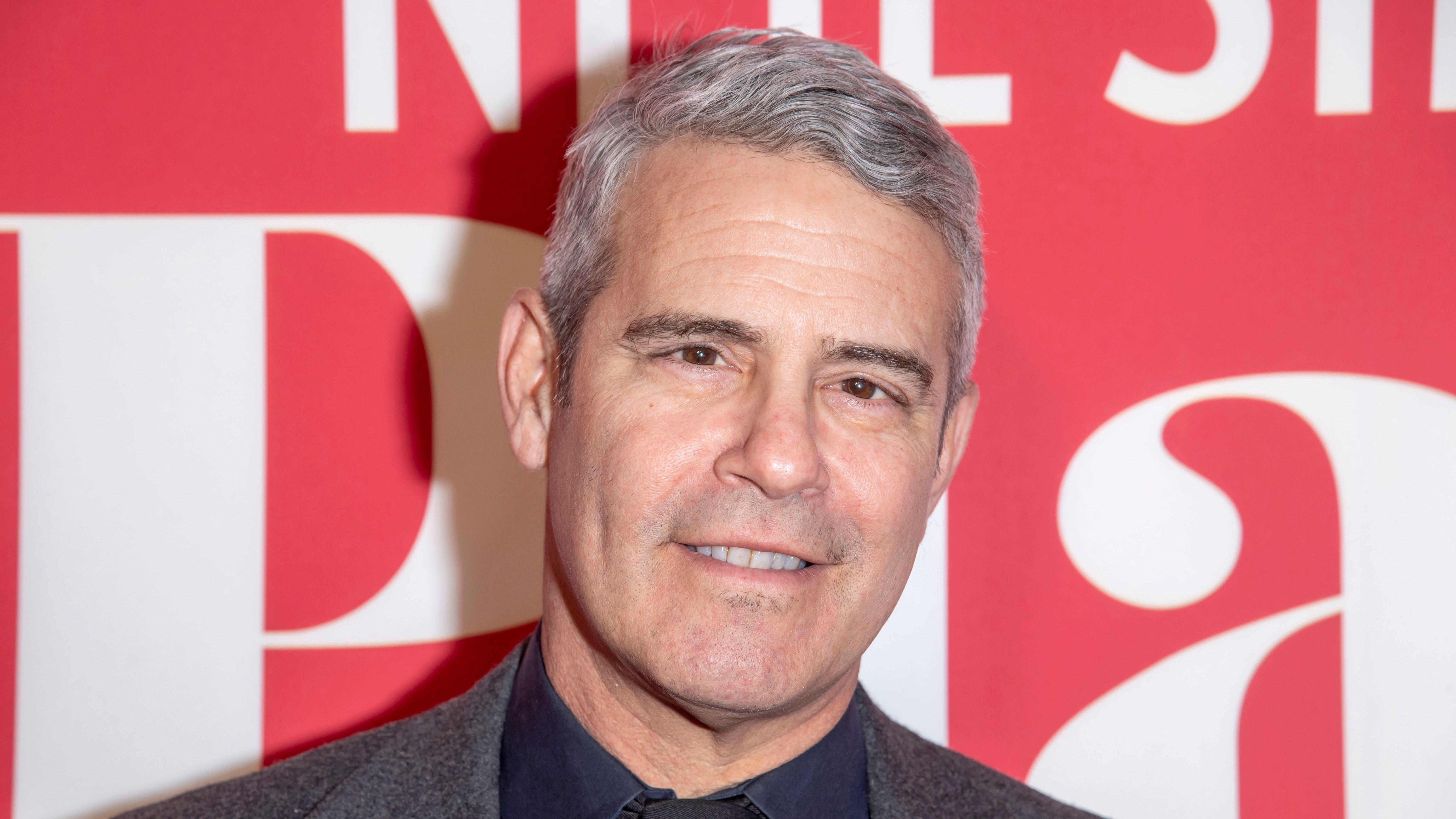 Andy Cohen smiles in navy shirt and gray blazer