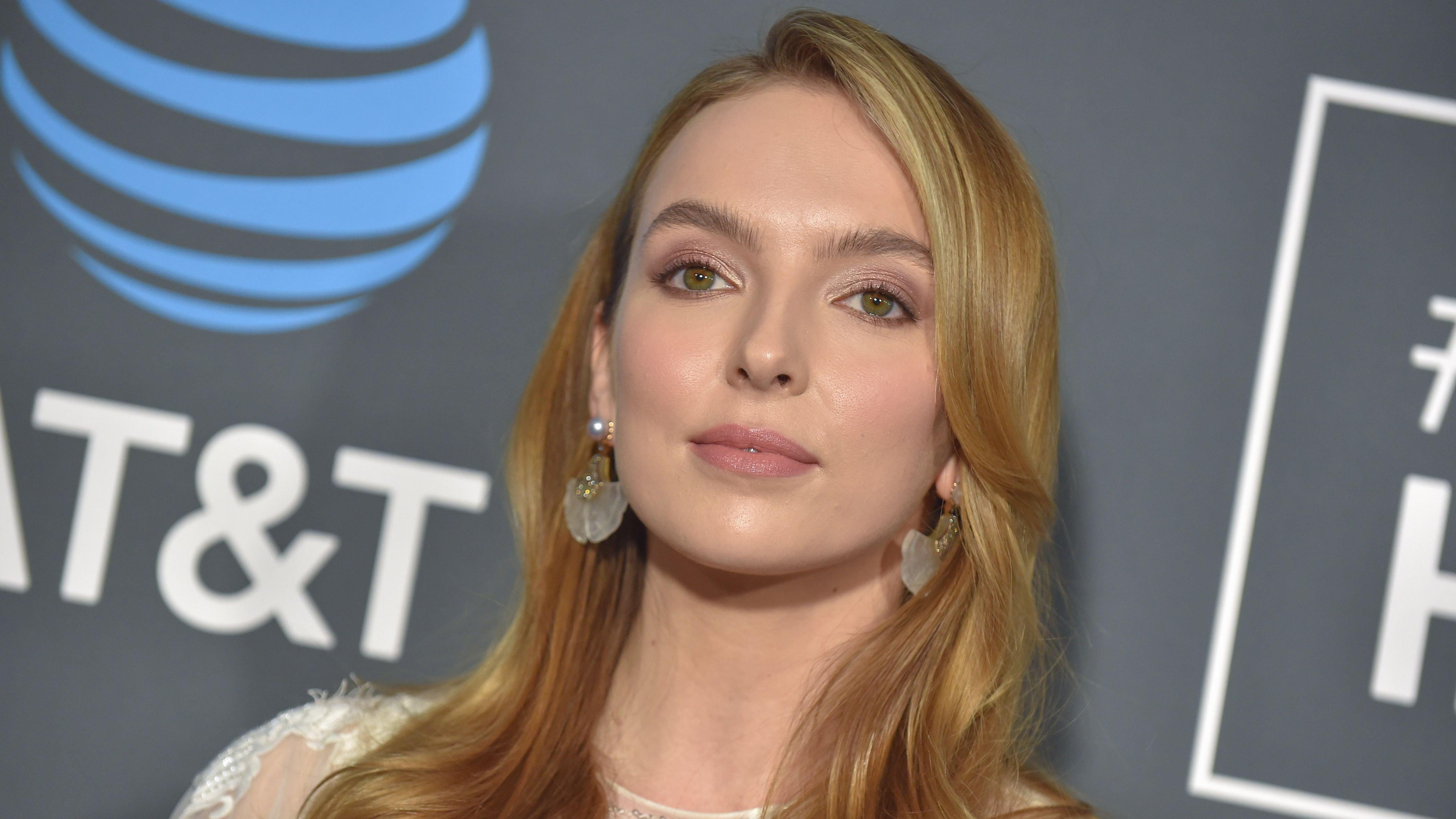 Jodie Comer attends an event sponsored by AT&T. She is wearing a white lace dress and large earrings. 