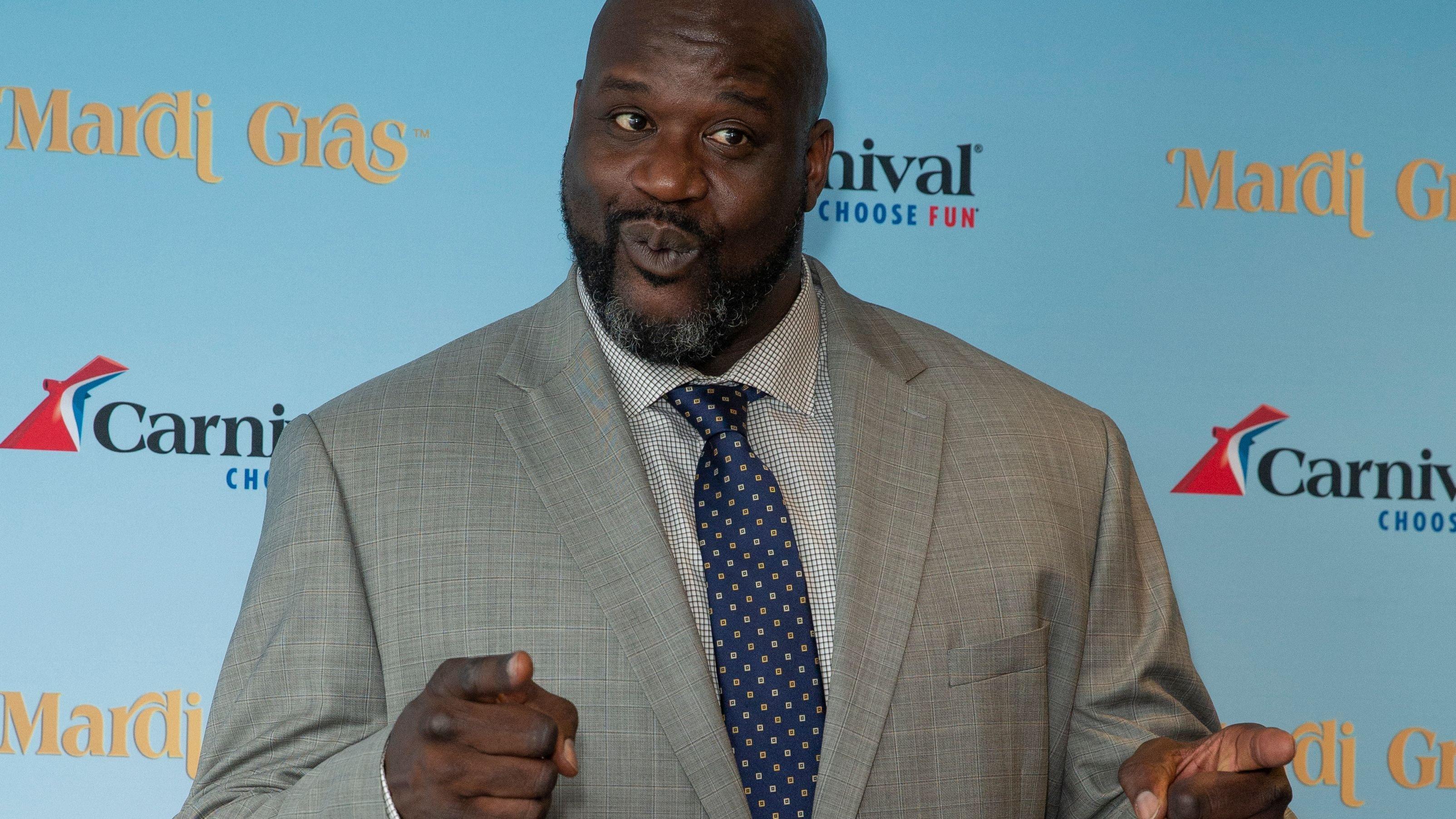 Shaquille O'Neal at an event in a grey suit pointing fingers at cameras